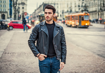 One handsome young man in urban setting in modern city, standing, wearing black leather jacket and jeans, looking away - Powered by Adobe