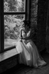 Young beautiful wedding bride in a beautiful long dress sitting in a chair in the apartment in the loft style near the window