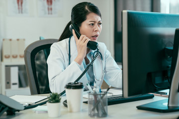 angry asian chinese woman doctor yells into phone. mad lady medical worker has conversation with patient bad emotions. confused young girl intern frowning talking looking at computer argue in clinic.