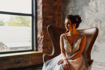 Young beautiful wedding bride in a beautiful long dress sitting in a chair in the apartment in the loft style near the window