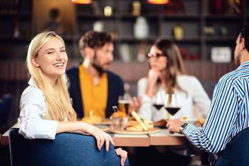 Young smiling Caucasian blonde cute woman dressed smart casual sitting at table in restaurant and looking over shoulder. In background are her friends having dinner.