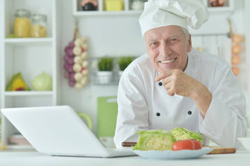 Portrait of elderly male chef cooking at table with laptop