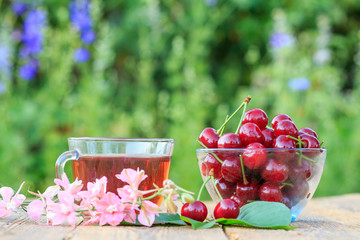 Red ripe cherry fruits with peduncles in glass bowl and cup of tea.
