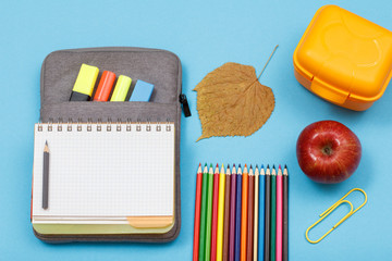Color pencils, apple, lunch box, dry leaf, open exercise book on bag-pencil case on blue background.