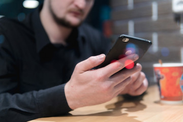 Man sits at table in cafe, night time, dark theme. Uses a smartphone and drinking coffee. Guy is chatting, working, blogging, learning online. Social media, marketing. Online education. window street.
