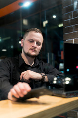 Business man looks at the phone, holds it in his hand in cafe. Night time view from the street. Dark theme, in black style. Guy is chatting, working, blogging. Social media. Online education. vertical