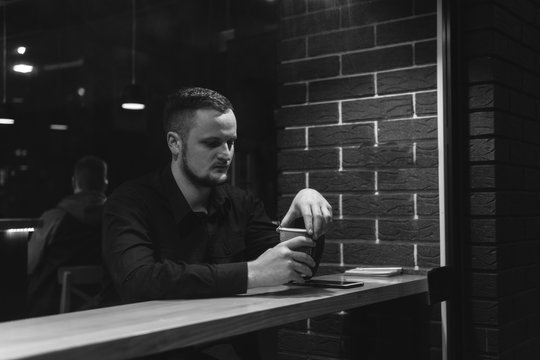 Man sits at table in cafe, night time, dark theme. black and white. alone drinking coffee. Guy is sad, dipression, thoughtfulness. Think. to plan. sadness to solve the problem. task. window street.