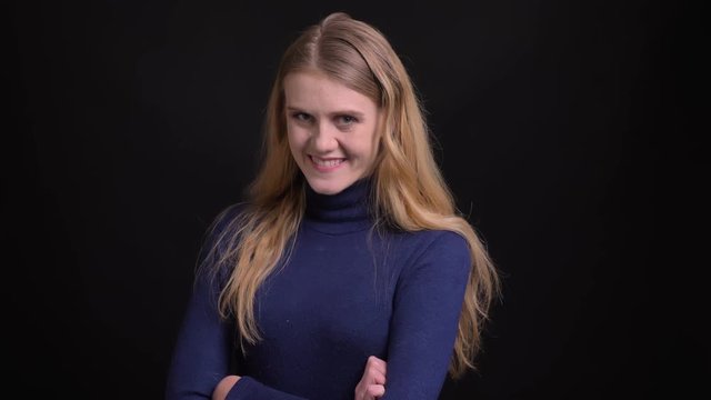 Young blonde model in blue sweater watching into camera with shy and pretty smile on black background.
