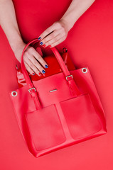 Bright summer fashion ladies accessories. Stylish red leather handbag or shopper bag on a red background. Women hands with bright manicure pulls a purse out of the bag. Top View. Flat Lay. Copy Space.