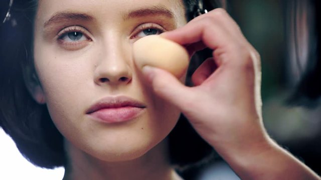 cropped view of makeup artist applying concealer under model eye with cosmetic sponge