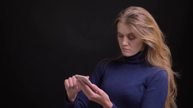Young blonde model swipes photos in smartphone and emotionally reacting on black background.
