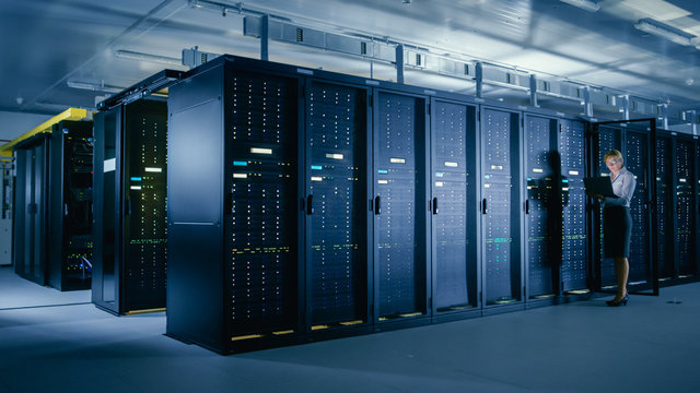 In Data Center: Female IT Technician Stands before Open Server Rack Cabinet, Uses Laptop Computer to Run Maintenance Diagnostics so that Mainframe Works at Optimal Functioning Level. 