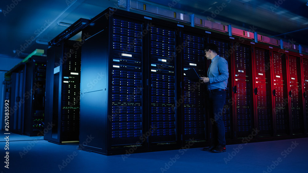 Poster in data center: male it specialist walks along the row of operational server racks, uses laptop for 