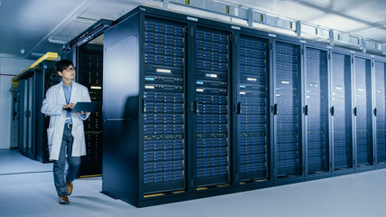 In Data Center: Male IT Specialist Wearing White Coat Walks Alongside Row of Server Racks, Uses...