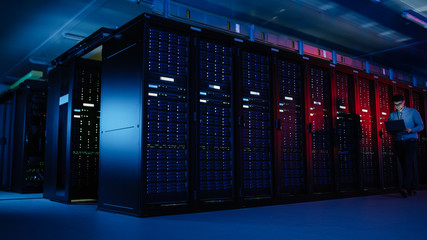 In Data Center: Male IT Specialist Walks along the Row of Operational Server Racks, Uses Laptop for Maintenance. Concept for Telecommunications, Cloud Computing, Artificial Intelligence, Supercomputer