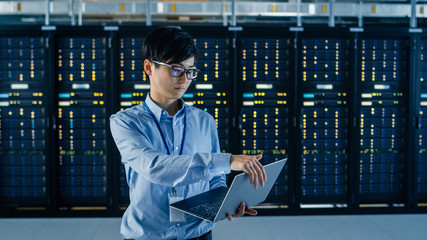 In Dark Data Center: Male IT Specialist Stands Beside Row of Operational Server Racks, Uses Laptop for Maintenance. Concept for Cloud Computing, Artificial Intelligence, Supercomputer, Cybersecurity