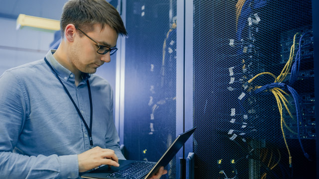 In Data Center IT Engineer Stands Before Working Server Rack Doing Routine Maintenance Check and Diagnostics Using Laptop. Visible Computer Hardware Equipment, Broadband Fiber Optic Cables LED Lights.