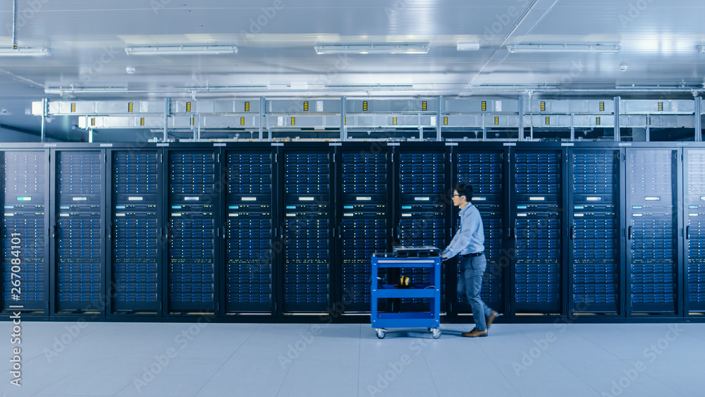 Fototapete In the Modern Data Center: IT Technician Working with Server Racks, Pushes Cart Between Rows of Server Racks. On a Pushcart New Hardware for System Update. Engineer Doing Maintenance and Diagnostics.
