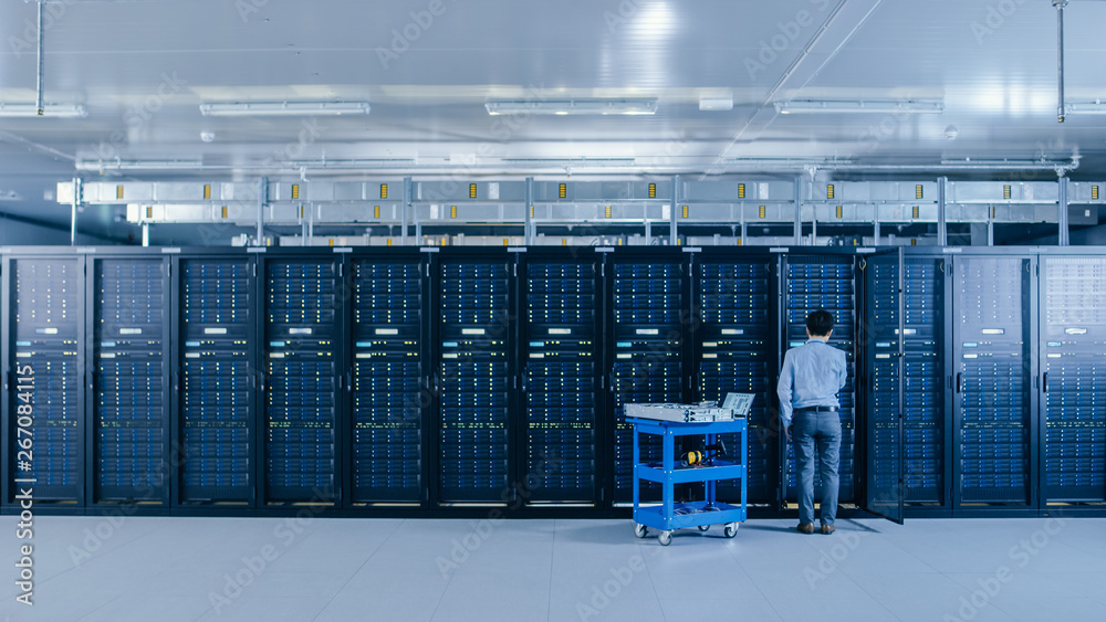 Aufkleber In the Data Center: IT Engineer Working with Server Racks Works with Opened Cabinet to Installs New Hardware for System Update, Cart Stands Beside the Row of Server Racks. Engineer Doing Maintenance.