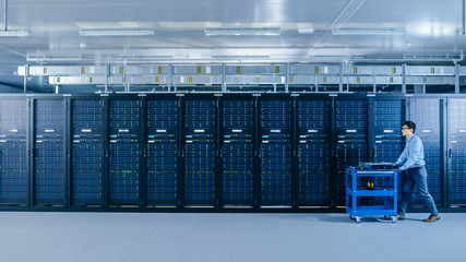 In the Modern Data Center: IT Technician Working with Server Racks, Pushes Cart Between Rows of...