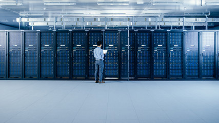 In the Modern Data Center: IT Engineer Standing Beside Open Server Rack Cabinets, Does Wireless...