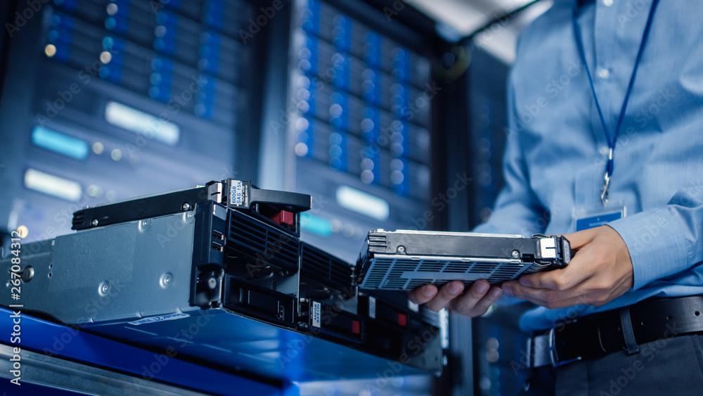 Poster In the Modern Data Center: IT Engineer is Holding New HDD Hard Drive Prepared for Installing Hardware Equipment into Server Rack. IT Specialist Doing Maintenance and Updating Hardware.