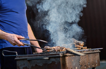 A man with tongs turns over lamb on a grill, on a sunny summer evening in nature, against the background of smoke