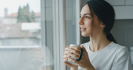 Portrait of a pensive young woman is drinking a tea and looking through the window in the morning in the kitchen.