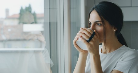 Portrait of a pensive young woman is drinking a tea and looking through the window in the morning...
