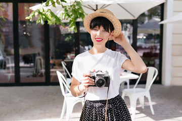 Pretty woman in elegant straw hat posing with charming smile and camera in hand on the street. Outdoor portrait of cute dark-haired girl in white shirt spending time in open-air cafe in morning.