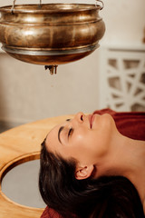 relaxed young woman lying under shirodhara vessel during ayurvedic procedure