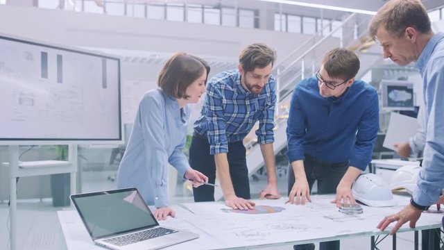 In the Industrial Engineering Facility: Diverse Group of Engineers and Technicians on a Meeting Gather Around Table with Engine Design Technical Drafts, Have Discussion, Analyse Technology