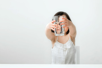 The little girl drinks water from a glass cup. The girl in a white suit drinks clear water. Girl shows us that she drinks water