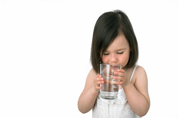 The little girl drinks water from a glass cup. The girl in a white suit drinks clear water. Girl shows us that she drinks water