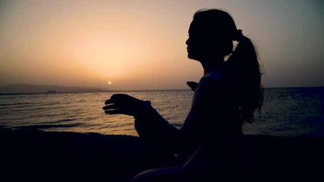 Sagoma di una bambina con i capelli a coda seduta sulla spiaggia mentre fa yoga di fronte al mare e al sole al tramonto.
