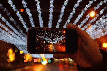 The hand with the phone making photos of lights, garlands and ornaments of the streets in the winter after rain in the night with reflection and Boke, Christmas, New Year.