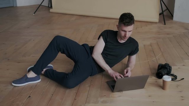 Stylish creative photographer retouching images on his laptop in comfort position while lying on the floor. He selecting illustrations for future working process and switches attention to the window.