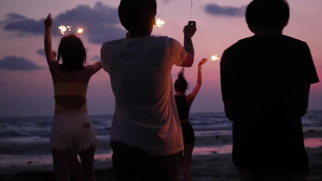 Asian teen group dancing and playing firework seaside together in beach summer with sunset background. Young asia happy emotion and anniversary celebration. 4K resolution and slow motion.