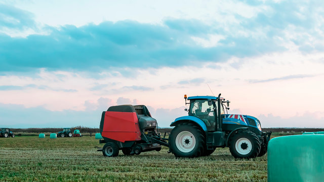 Agricultural Work In A Field At Sunset. Equipment For Forage. Film Wrapping System. Round Bales Of Feed For Farm Animals.