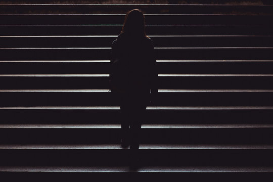 Black silhouette of a young woman casually dressed climbing up the city street stairs – Single person walking in the dark of the night – Concept image for leadership, achievement or independence