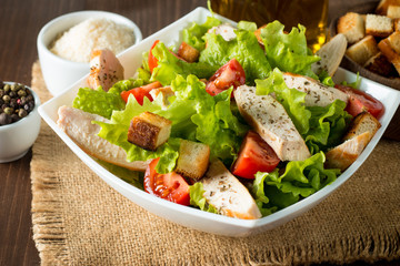 Fresh salad made of tomato, ruccola, chicken breast, eggs, arugula, crackers and spices. Caesar salad in a white, transparent bowl on wooden background