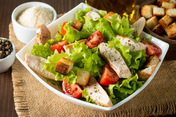 Fresh salad made of tomato, ruccola, chicken breast, eggs, arugula, crackers and spices. Caesar salad in a white, transparent bowl on wooden background