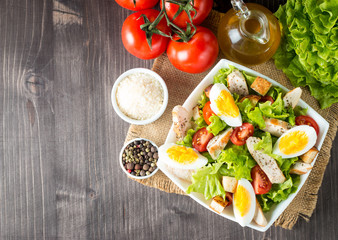 Fresh salad made of tomato, ruccola, chicken breast, eggs, arugula, crackers and spices. Caesar salad in a white, transparent bowl on wooden background