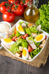 Fresh salad made of tomato, ruccola, chicken breast, eggs, arugula, crackers and spices. Caesar salad in a white, transparent bowl on wooden background