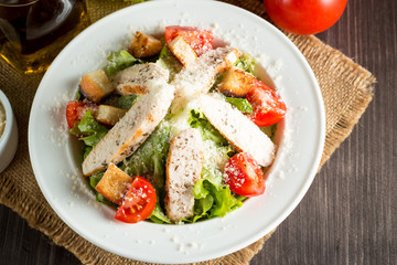 Fresh salad made of tomato, ruccola, chicken breast, eggs, arugula, crackers and spices. Caesar salad in a white, transparent bowl on wooden background