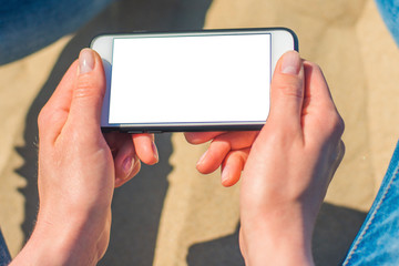 Image Mockup of beautiful hands of a woman holding a white mobile phone with a blank screen on her hip on the beach, on the sand. Close up.