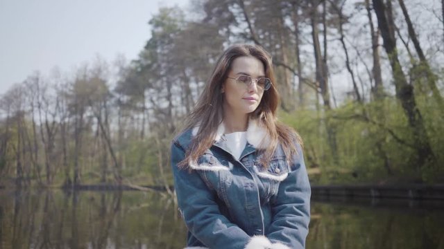 Portrait of a graceful young woman in sunglasses and a denim jacket floating on a boat on a lake or river. Beautiful brunette is actively relaxing on a day off or traveling enjoying nature.