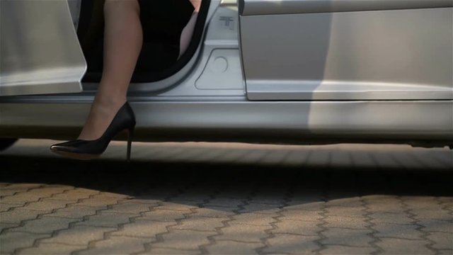 Close-Up Of Young Lady In Spotted Dress Getting Out Of Car And Walks By Road. Low Angle View With Focus On Woman's Legs.
