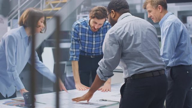 In the Industrial Engineering Facility: Diverse Group of Engineers and Technicians on a Meeting Gather Around Table Unravel Sheets of Engine Design Technical Drafts, Analyse Drawings. High Angle