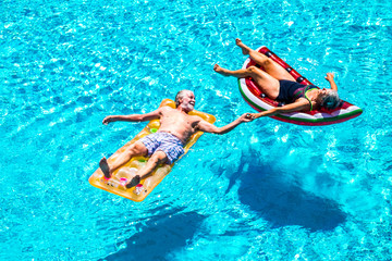 Obraz na płótnie Canvas Happy relaxed people caucasian senior adult couple taking hands lay down with trendy coloured lilos on a blue water swimming pool during a summer holiday vacation - enjoying active retired lifestyle